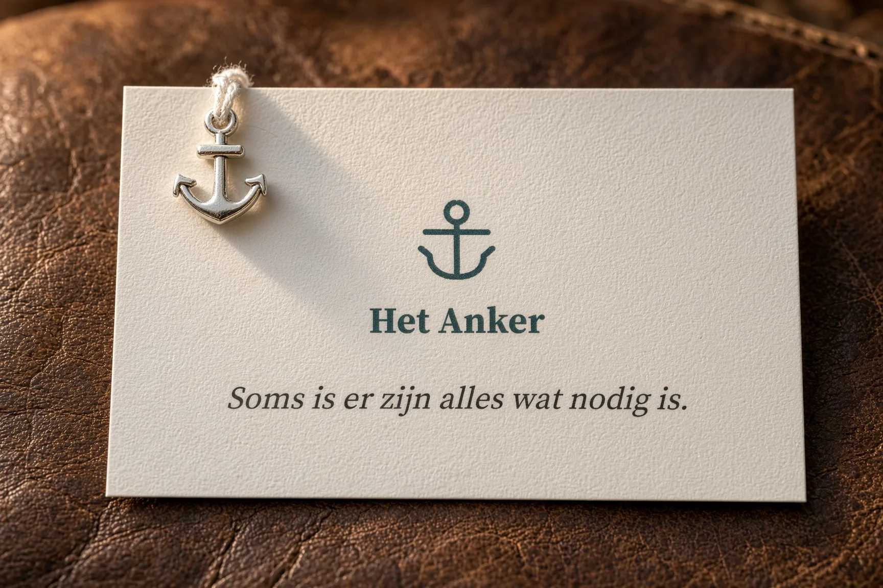 Sfeerbeeld: een crèmekleurig kaartje met het ankerlogo, de naam Het Anker en de tekst «Soms is er zijn alles wat nodig is», met een klein zilveren anker aan een touwtje, op bruin leer.